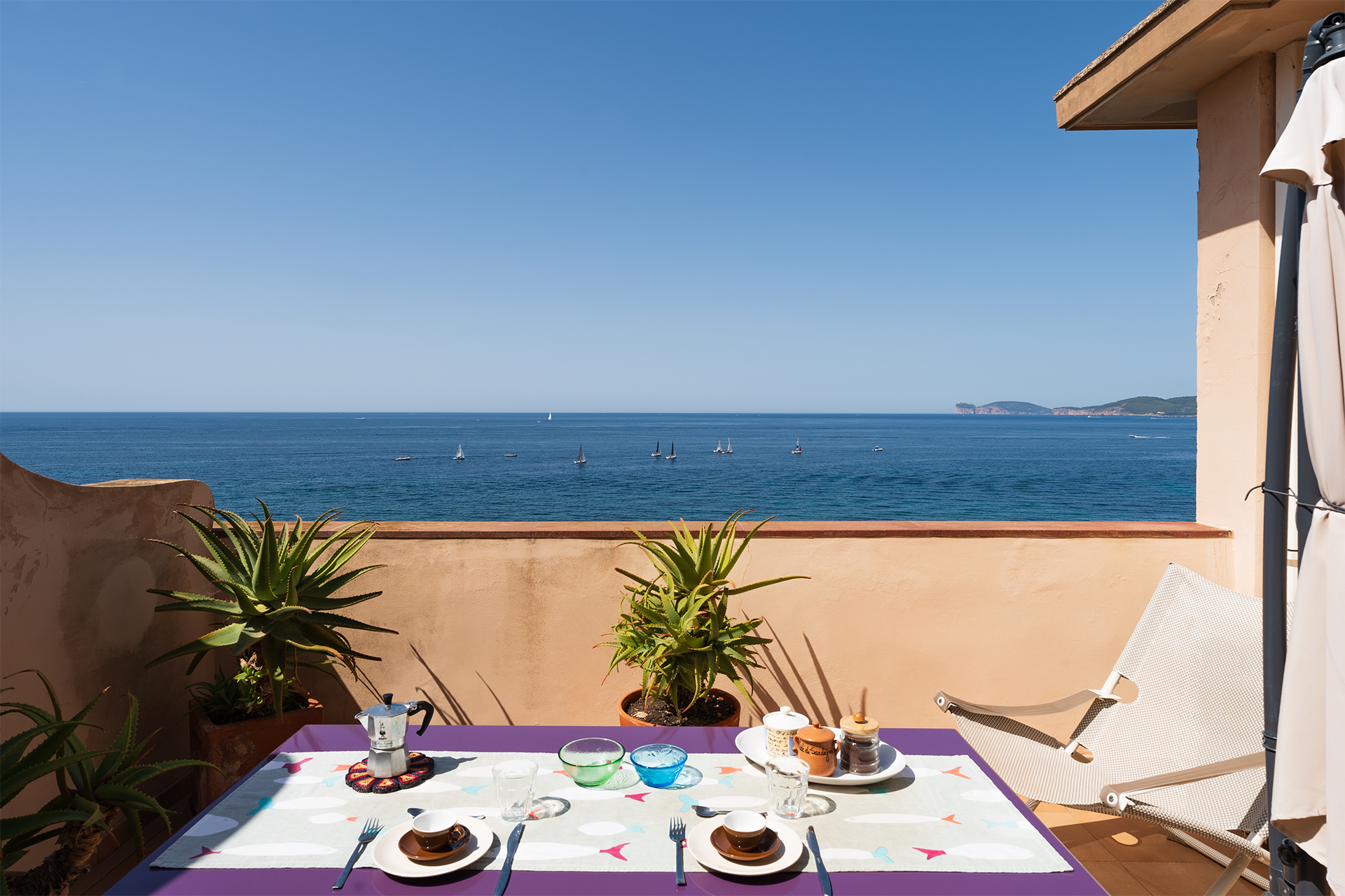 Terrazza panoramica con tavolo, seduta relax e barche a vela sul mare di Alghero